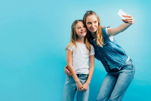 Madre in denim che prende selfie su smartphone con bambino che indossa jeans e t-shirt bianca isolata su blu — Foto stock