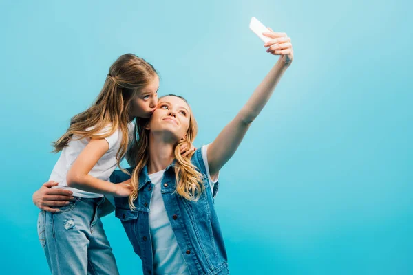 Criança em camiseta branca e jeans beijando mãe em colete jeans levando selfie no smartphone isolado em azul — Fotografia de Stock