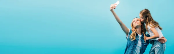 Imagen horizontal del niño besando a la madre tomando selfie en el teléfono inteligente aislado en azul - foto de stock