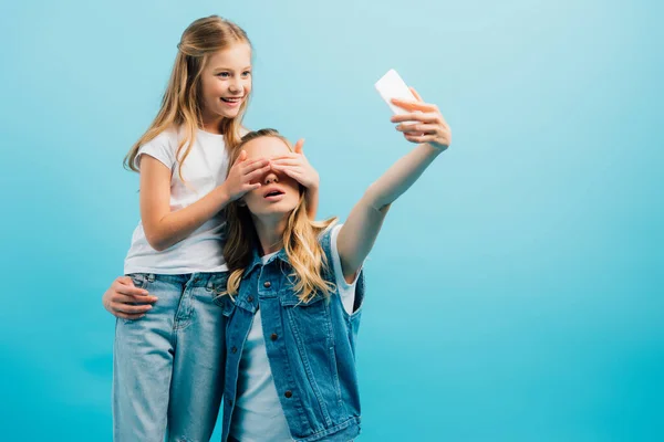 Jovem mulher tomando selfie no smartphone enquanto filha cobrindo seu rosto com as mãos isoladas no azul — Fotografia de Stock