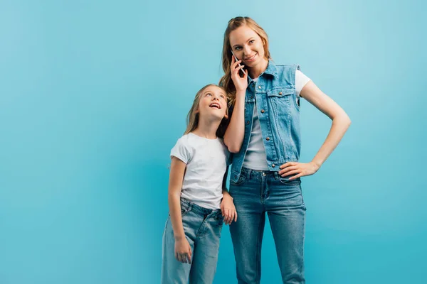 Молода жінка в джинсовому одязі розмовляє на смартфоні, стоячи з рукою на стегні біля допитливої дочки ізольовано на синьому — стокове фото