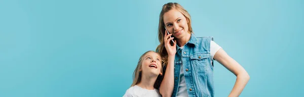 Tiro panorámico de mujer joven en chaleco de mezclilla hablando en el teléfono inteligente cerca de hija curiosa aislada en azul - foto de stock