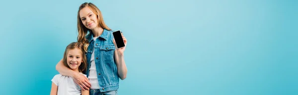 Immagine orizzontale di madre in gilet di denim che mostra smartphone con schermo bianco mentre abbraccia figlia isolata su blu — Foto stock