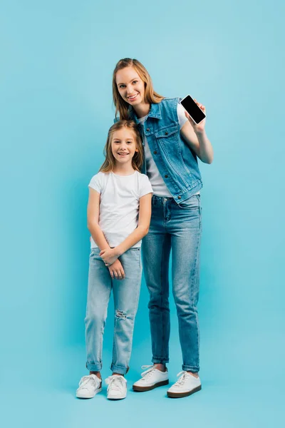 Full length view of mother in denim clothes showing smartphone with blank screen near daughter on blue — Stock Photo