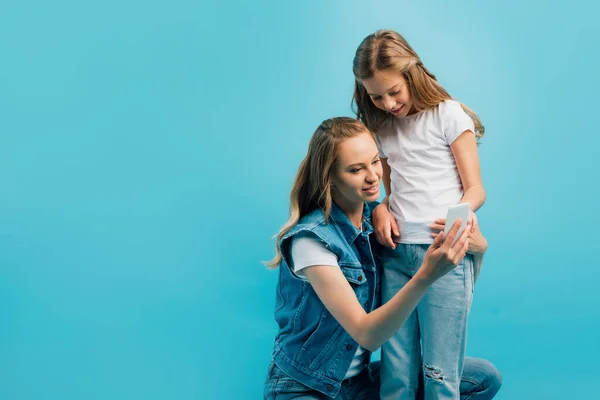 Mujer joven en chaleco de mezclilla usando un teléfono inteligente mientras se agacha cerca de la hija aislada en azul - foto de stock