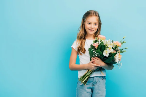 Kind in weißem T-Shirt und blauer Jeans blickt in die Kamera, während es Blumenstrauß isoliert auf blauem Grund hält — Stockfoto