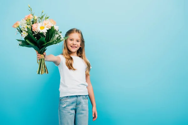 Niña en camiseta blanca y jeans con ramo de flores en la mano extendida mientras mira a la cámara aislada en azul - foto de stock