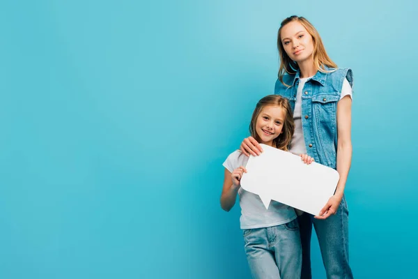 Giovane madre in denim vestiti tenendo discorso bolla con figlia isolata su blu — Foto stock