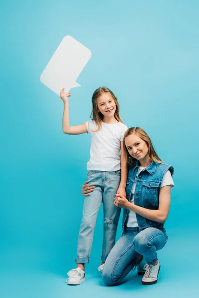 Молодая женщина в джинсовой одежде приседает рядом с дочерью в белой футболке и джинсах, держа речевой пузырь на синем — стоковое фото