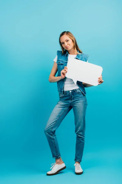 Вид в полный рост женщины в джинсовом жилете и джинсах, держащей пузырь речи на голубом фоне — стоковое фото