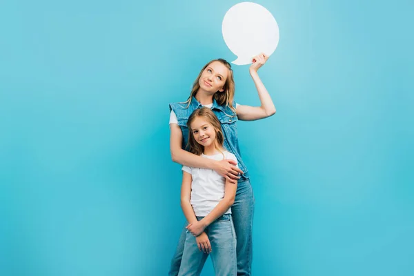 Chica en camiseta blanca y jeans mirando a la cámara mientras la madre sostiene la burbuja de pensamiento aislado en azul - foto de stock