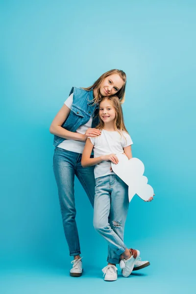 Вид в полный рост на молодую женщину в джинсовой одежде, трогающую дочь, держащую мыльный пузырь на голубом фоне — стоковое фото
