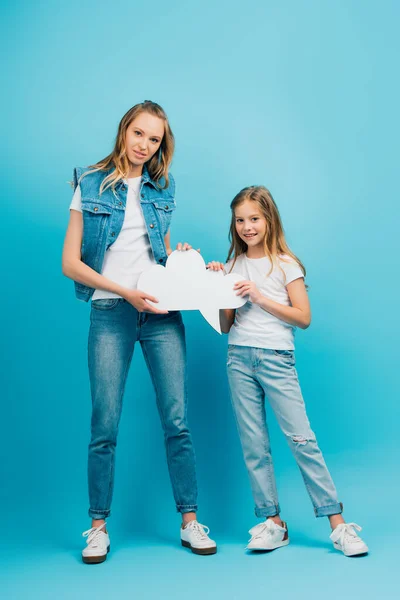 Вид в полный рост матери и дочери в джинсовой одежде, держащей мыльный пузырь, глядя в камеру на голубом фоне — стоковое фото