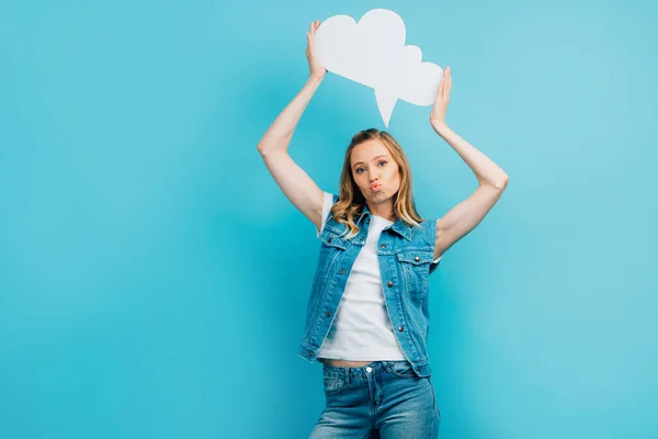 Young woman in denim vest pouting lips while holding thought bubble above head isolated on blue — Stock Photo