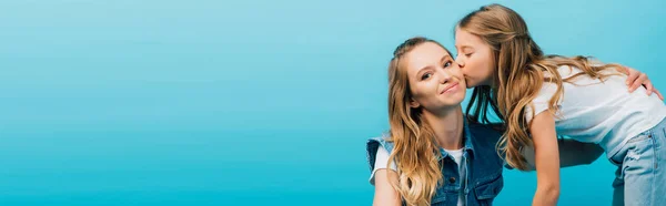 Imagem horizontal da jovem mulher olhando para a câmera enquanto filha beijando-a isolada no azul — Fotografia de Stock