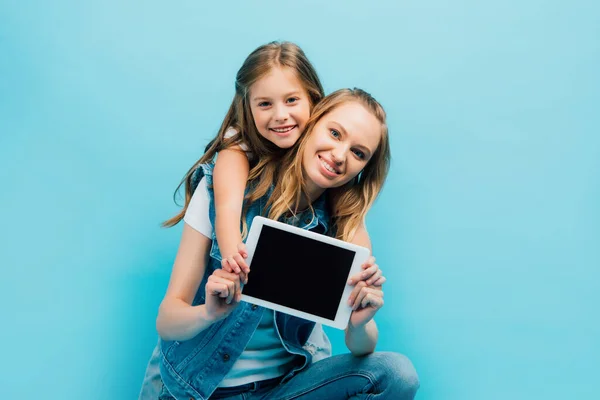 Mutter und Kind in Jeanskleidung zeigen ein digitales Tablet mit leerem Bildschirm, während sie die Kamera isoliert auf blauem Grund betrachten — Stockfoto