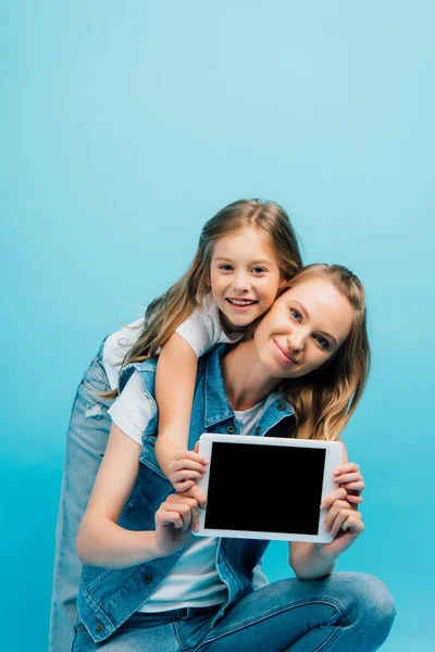Мать и дочь в джинсовой одежде показывая цифровой планшет с чистым экраном и глядя на камеру изолированы на синий — стоковое фото