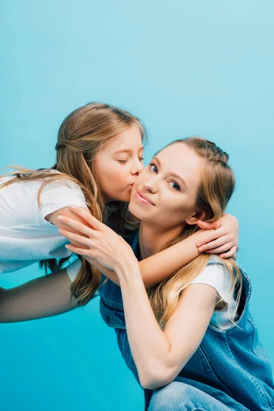 Молодая женщина смотрит в камеру, в то время как дочь целует ее с закрытыми глазами изолированы на синий — стоковое фото