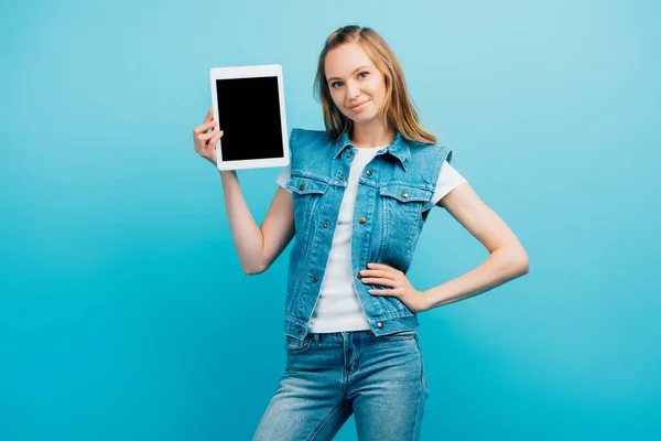 Woman in denim clothes standing with hand on hip and showing digital tablet with blank screen isolated on blue — Stock Photo