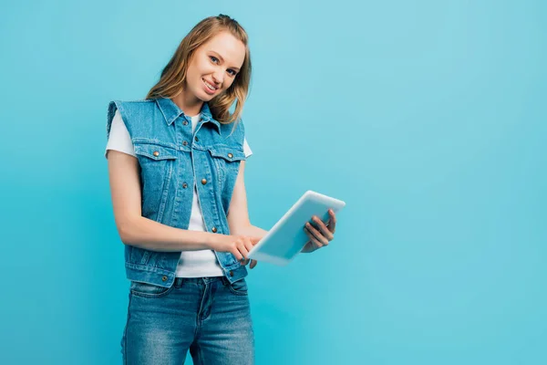 Young woman in denim clothes using digital tablet while looking at camera isolated on blue — Stock Photo