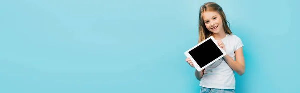 Imagem horizontal da menina em camiseta branca mostrando tablet digital com tela em branco enquanto olha para a câmera isolada em azul — Fotografia de Stock