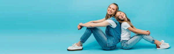 Panoramic concept of young woman with daughter in denim clothes sitting back to back on blue — Stock Photo