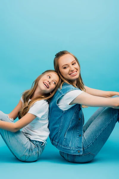 Madre e figlia in denim vestiti seduti sul pavimento schiena a schiena su blu — Foto stock