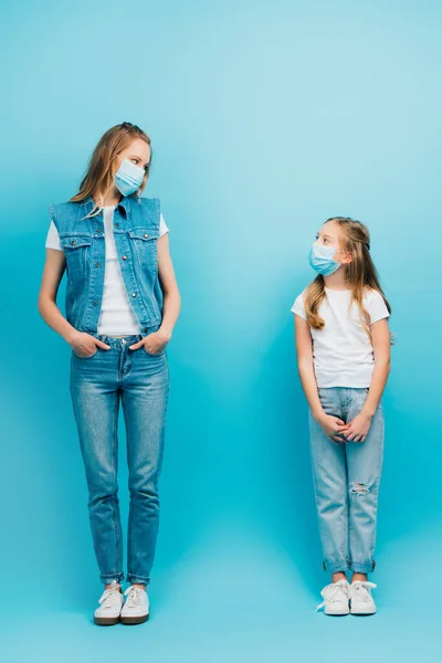 Visão de comprimento total de mulher e filha em roupas jeans e máscaras médicas olhando um para o outro, enquanto em pé no azul — Fotografia de Stock