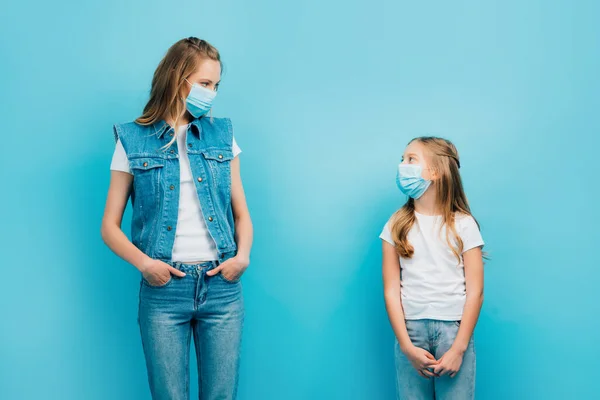 Mãe e filho em roupas jeans e máscaras médicas olhando um para o outro, enquanto em pé isolado em azul — Fotografia de Stock