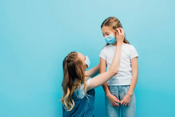 Joven mujer fijación máscara protectora en la cara de la hija aislado en azul - foto de stock