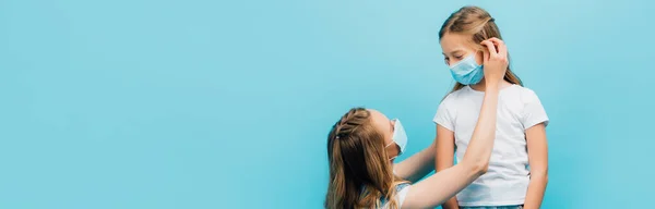 Horizontal image of mother putting on medical mask on daughter isolated on blue — Stock Photo