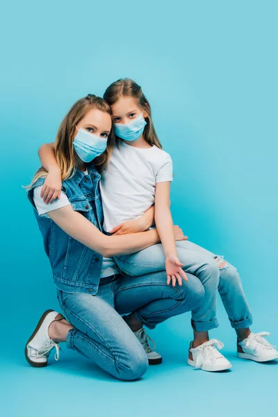 Chica en camiseta blanca sentada en el regazo de la madre en ropa de mezclilla mientras usa máscaras médicas y mirando a la cámara en azul - foto de stock