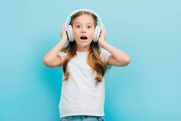 Excitada chica en camiseta blanca tocando auriculares inalámbricos mientras mira a la cámara aislada en azul - foto de stock