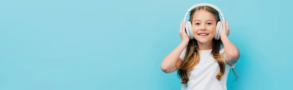 Horizontal image of child in white t-shirt touching wireless headphones isolated on blue — Stock Photo