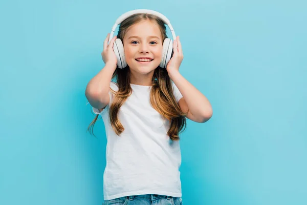 Chica en camiseta blanca tocando auriculares inalámbricos mientras mira la cámara aislada en azul - foto de stock