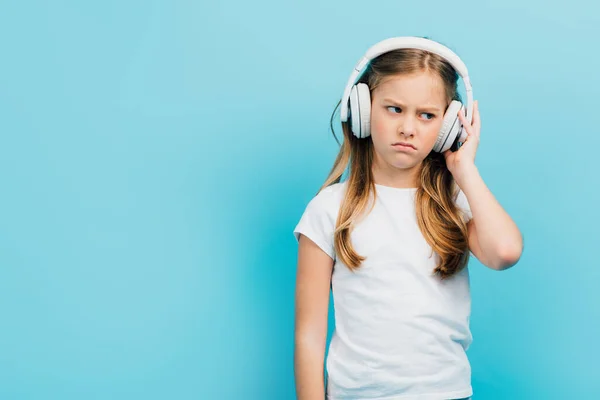 Chica disgustada en camiseta blanca tocando auriculares inalámbricos aislados en azul - foto de stock