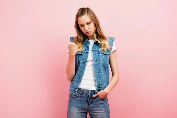 Irritated woman in denim clothes showing clenched fist while standing with hand in pocket isolated on pink — Stock Photo