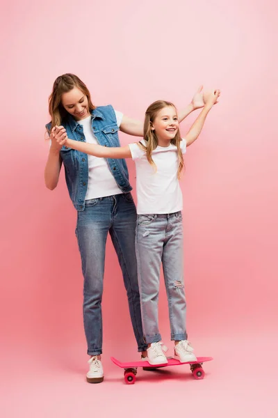 Full length view of woman in denim clothes supporting daughter standing on penny board by holding her hands on pink — Stock Photo