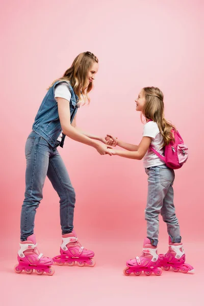 Мать и дочь в джинсовой одежде держатся за руки и смотрят друг на друга, стоя на роликовых коньках вместе на розовом — стоковое фото