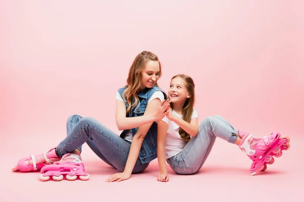 Mother and daughter in denim clothes sitting on floor in roller skates and giving high five on pink — Stock Photo