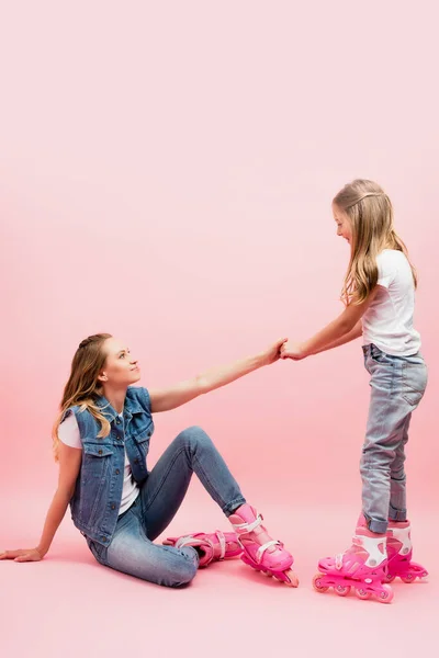 Hija cogida de la mano de la madre caída sentada en el suelo en patines sobre rosa - foto de stock
