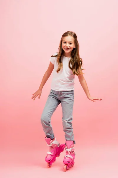 Aufgeregtes Mädchen in weißem T-Shirt, Jeans und Rollschuhen blickt in die Kamera auf rosa — Stockfoto