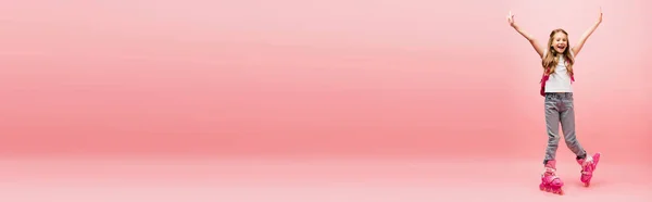 Горизонтальне зображення збудженої дитини в білій футболці, джинсах і ковзанах, дивлячись на камеру з піднятими руками на рожевому — стокове фото