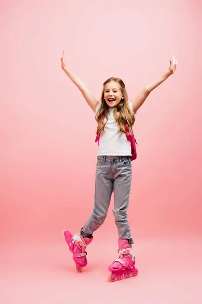 Excited girl in white t-shirt, jeans and rolling skates looking at camera with raised hands on pink — Stock Photo