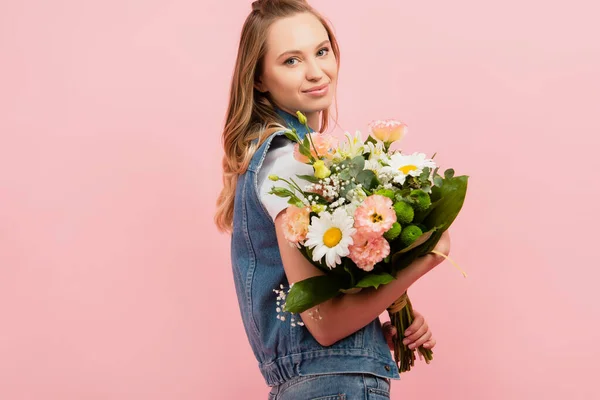 Junge Frau in Jeanskleidung hält Blumenstrauß in der Hand und blickt vereinzelt in die Kamera auf rosa — Stockfoto