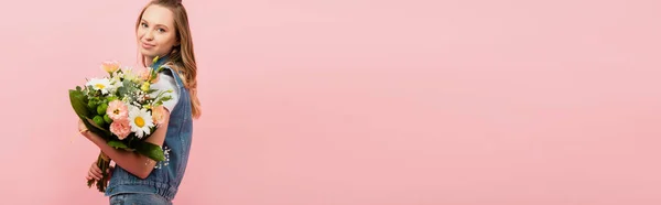 Horizontales Bild einer jungen Frau in Jeansweste mit Blumenstrauß und Blick in die Kamera isoliert auf rosa — Stockfoto