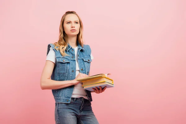 Вдумлива молода жінка в джинсовому жилеті, дивлячись далеко, тримаючи книги ізольовані на рожевому — стокове фото