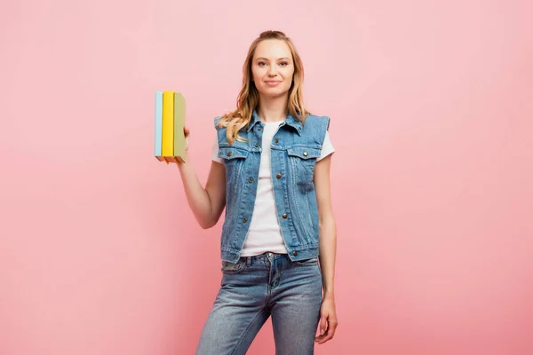 Молода жінка в джинсовому жилеті і джинсах тримає книги ізольовані на рожевому — стокове фото