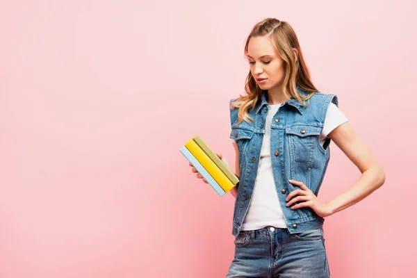 Donna seria in denim che tiene libri in piedi con mano sull'anca isolata su rosa — Foto stock