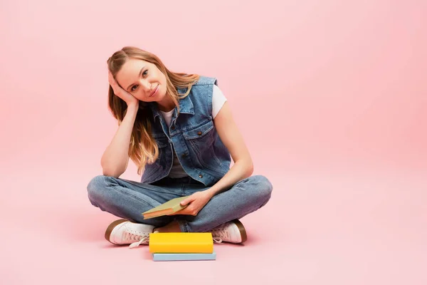 Jeune femme en gilet denim et jeans assis sur le sol avec les jambes croisées près des livres sur rose — Photo de stock